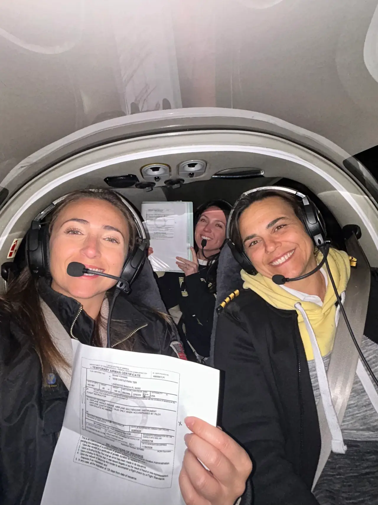 Women pilots with headsets in plane cockpit holding certificates and smiling while looking at the camera